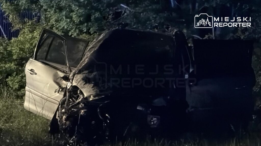 Zniszczony samochód osobowy stoi na poboczu drogi, otoczony gęstą roślinnością.