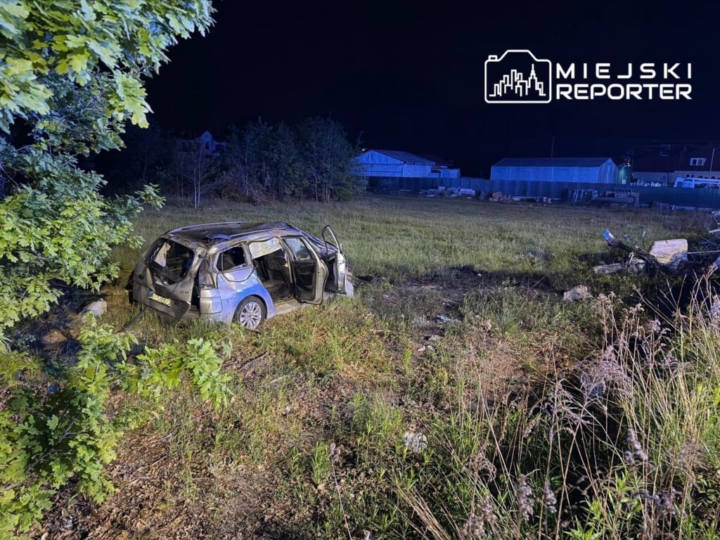 Uszkodzony samochód osobowy stoi na trawie w ciemnym otoczeniu, obok widoczne budynki i roślinność.