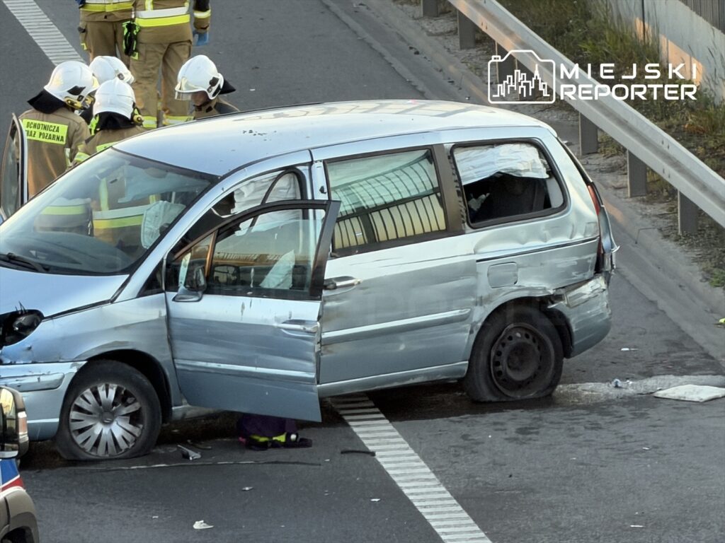 W miejscu wypadku stoi uszkodzony niebieski minivan z otwartymi drzwiami, obok grupka strażaków.