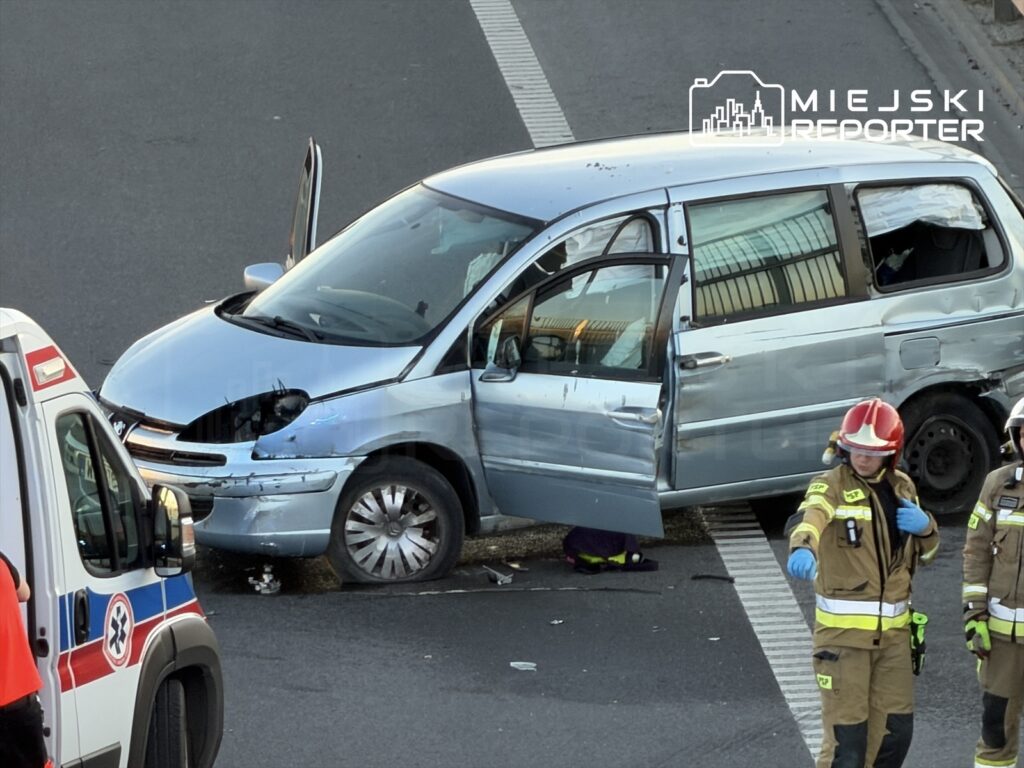 Wypadek drogowy z udziałem uszkodzonego niebieskiego minivana, obok pracują strażacy i zespół ratunkowy.