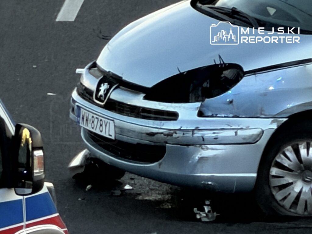 Uszkodzony samochód osobowy marki Peugeot z wgniecionym przodem na jezdni obok radiowozu policyjnego.