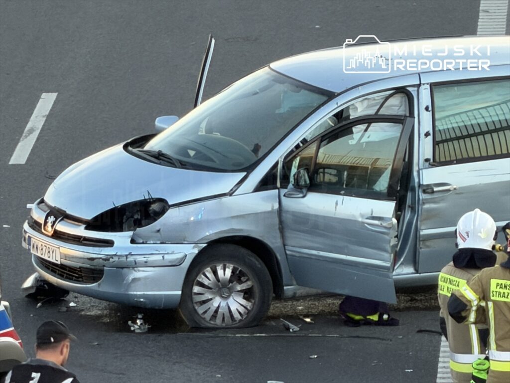 Srebrny samochód osobowy z uszkodzoną karoserią stoi na jezdni, obok znajdują się strażacy w mundurach.