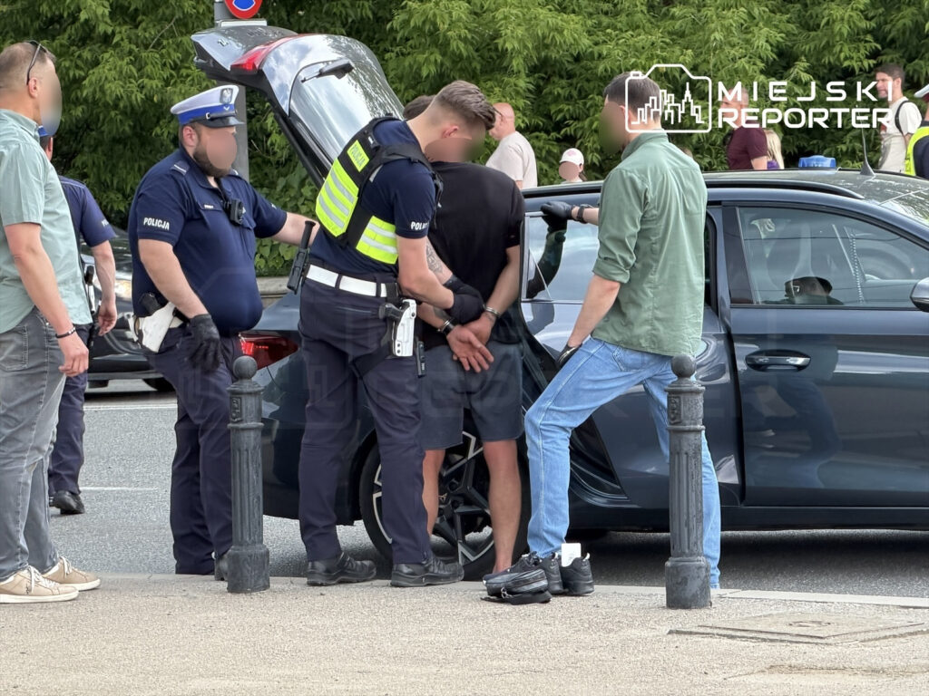 Policjanci zatrzymują mężczyznę z rękami skrępowanymi za plecami obok otwartego bagażnika samochodu.