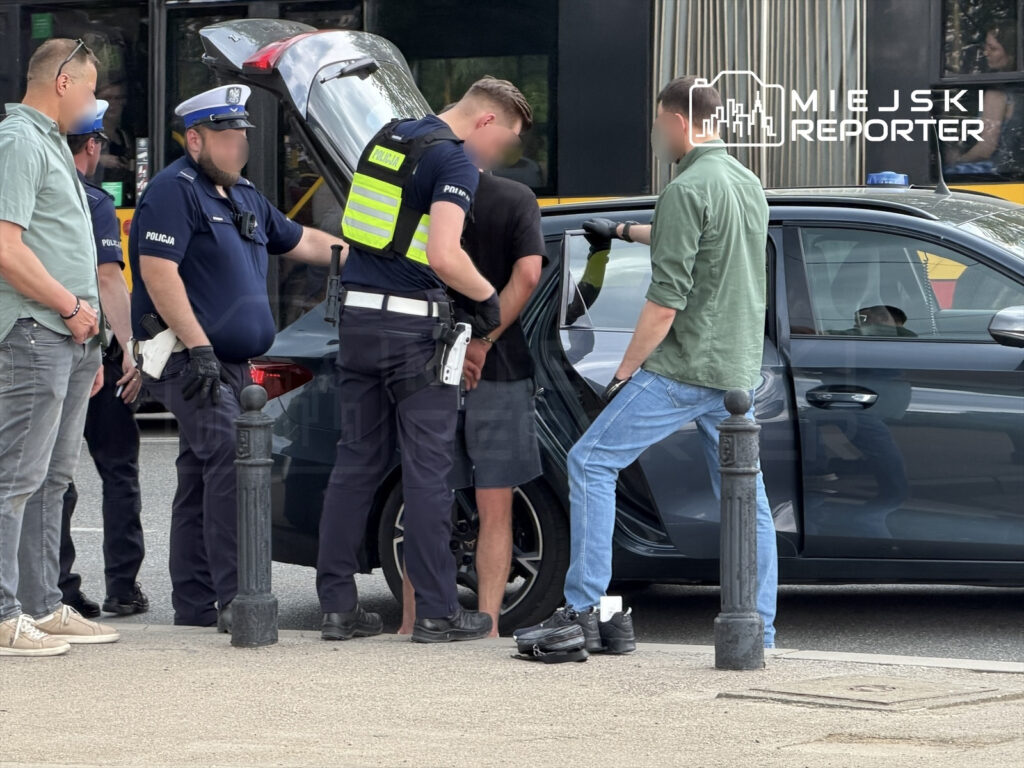 Grupa policjantów prowadzi kontrolę mężczyzny przy otwartym bagażniku samochodu na ulicy.