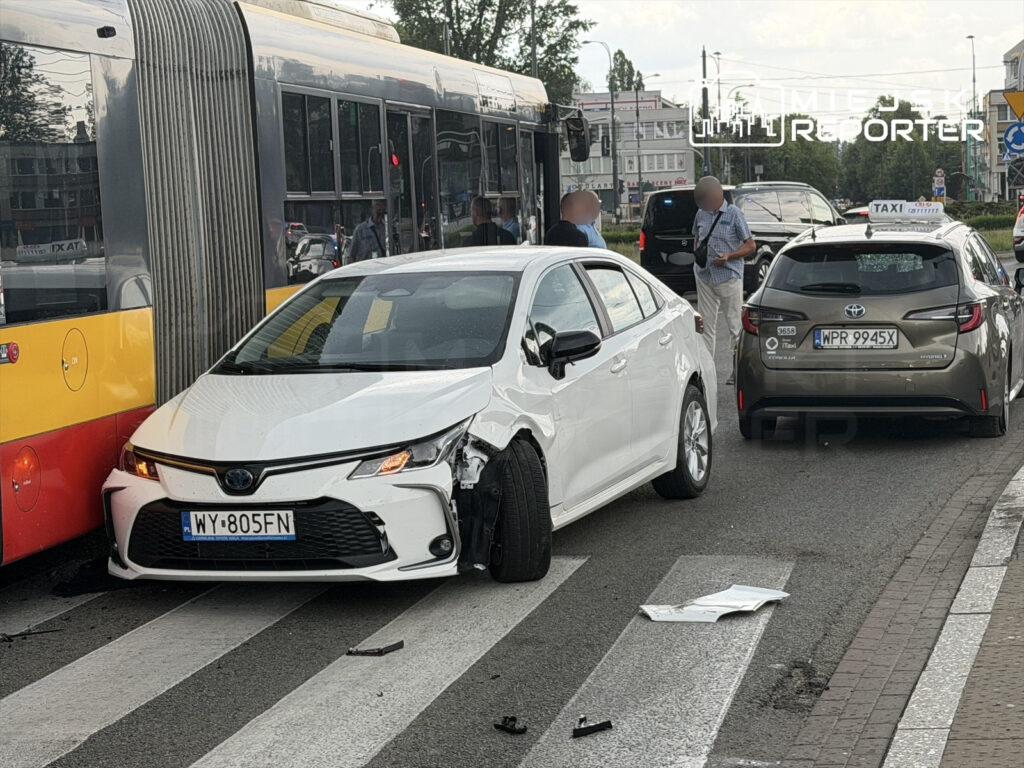 Biały samochód osobowy z uszkodzoną przednią częścią stoi obok tramwaju na przejściu dla pieszych, a mężczyzna rozmawia z kierowcą taksówki.