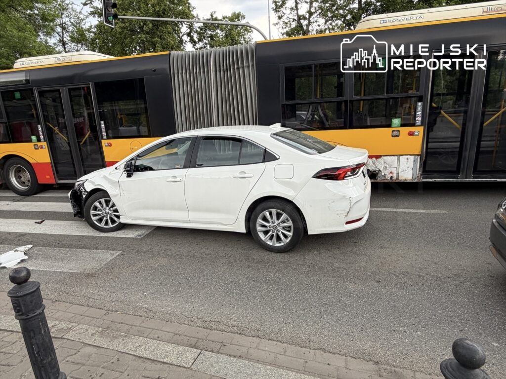 Biały samochód osobowy z uszkodzoną lewą stroną stoi na przejściu dla pieszych obok żółtego autobusu komunikacji miejskiej.