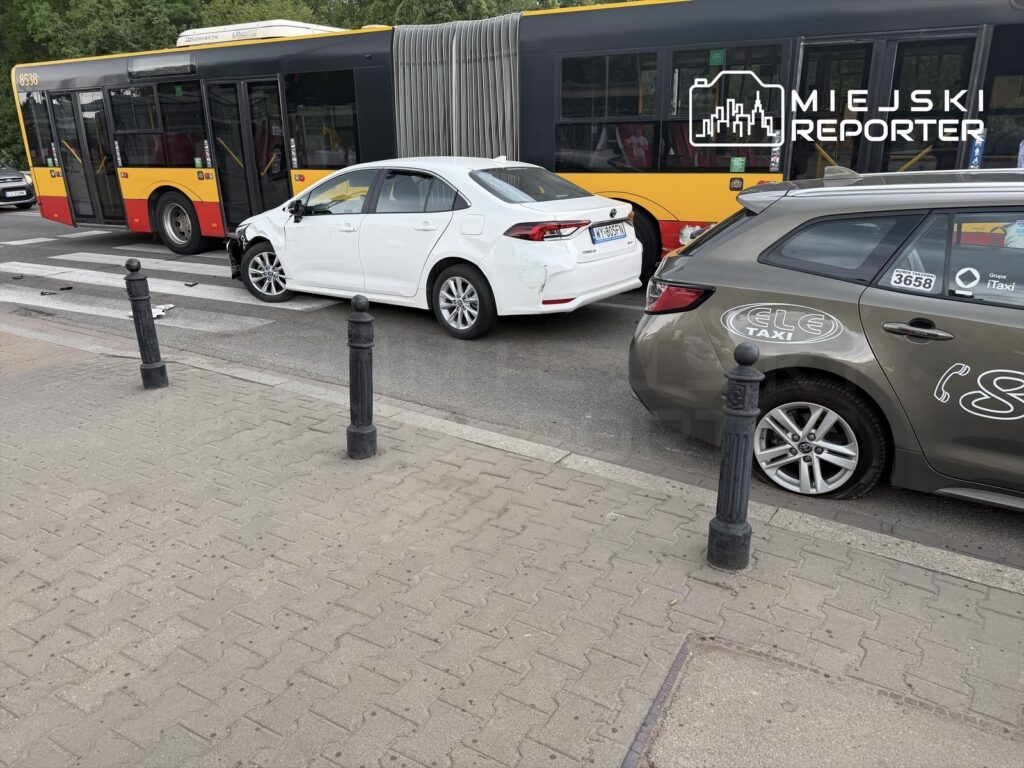 Biały samochód osobowy z uszkodzeniami zaparkowany obok autobusu miejskiego na przejściu dla pieszych.