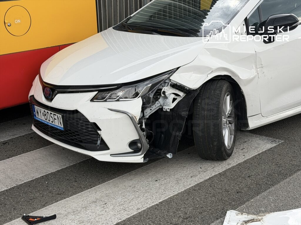 Uszkodzony biały samochód osobowy zatrzymany na pasach, obok żółtego autobusu. Widoczna uszkodzona przednia część pojazdu.