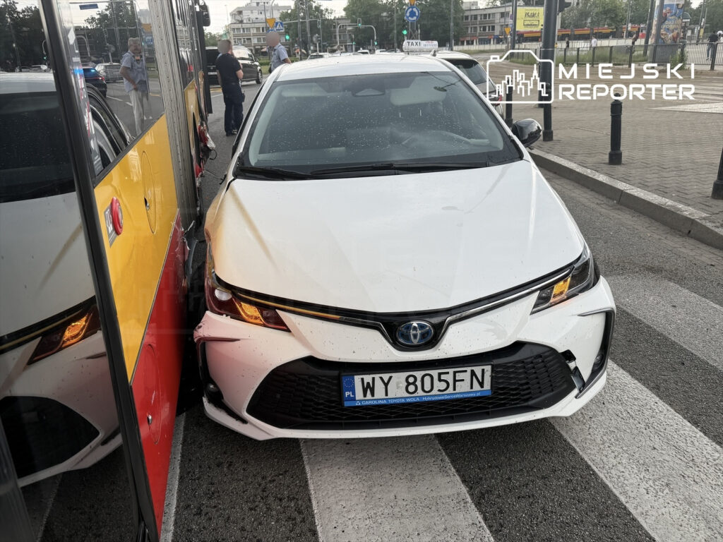 Biały samochód osobowy zderzony z żółtym autobusem na przystanku, w tle przechodnie i sygnalizacja świetlna.