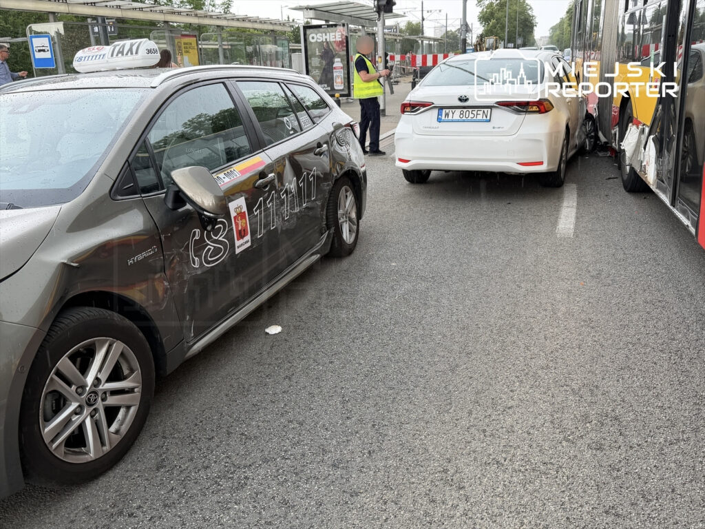 W zderzeniu uczestniczy taksówka z uszkodzoną boczną częścią oraz samochód osobowy z widocznymi obrażeniami tyłu, obok stoi pracownik w kamizelce odblaskowej.