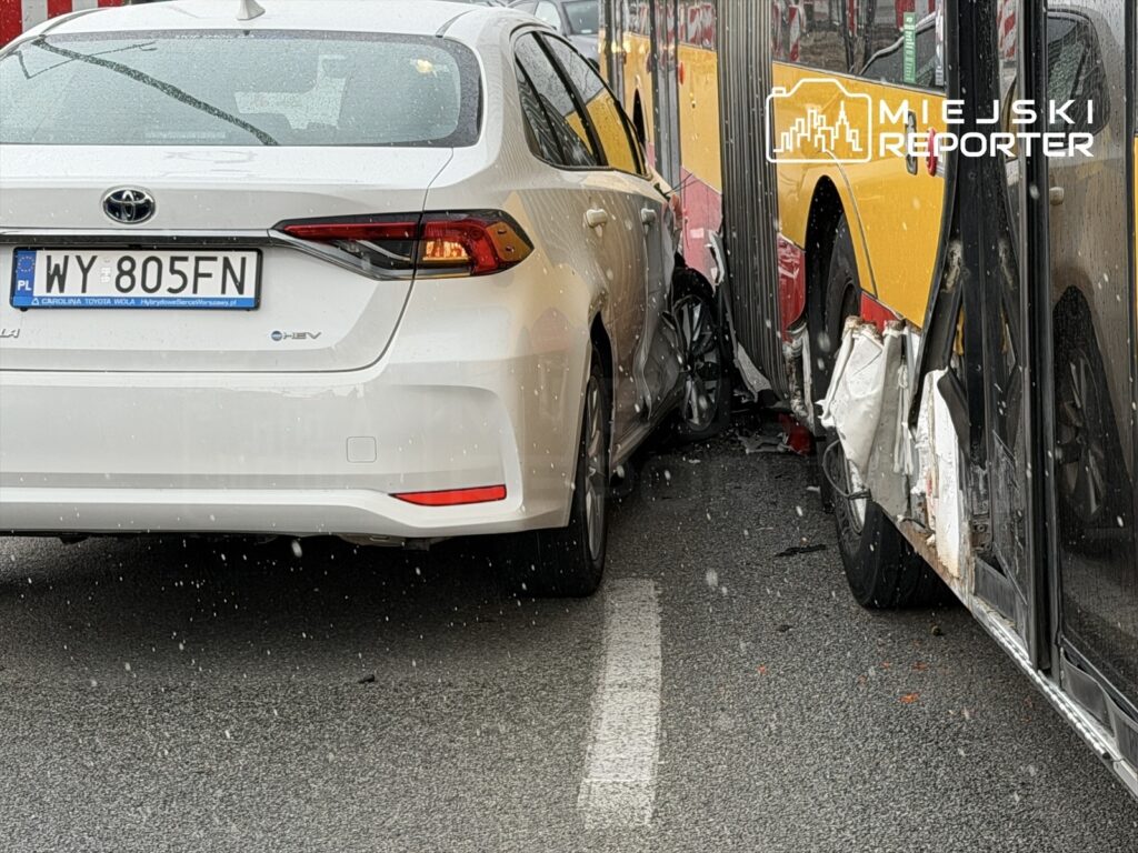 Biały samochód osobowy zderzony z czerwono-żółtym autobusem na mokrej nawierzchni drogi.