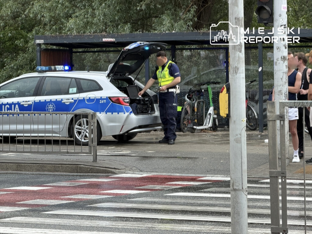 Policjant w mundurze sprawdzający bagażnik radiowozu przy przystanku autobusowym, w tle rowery i piesi.