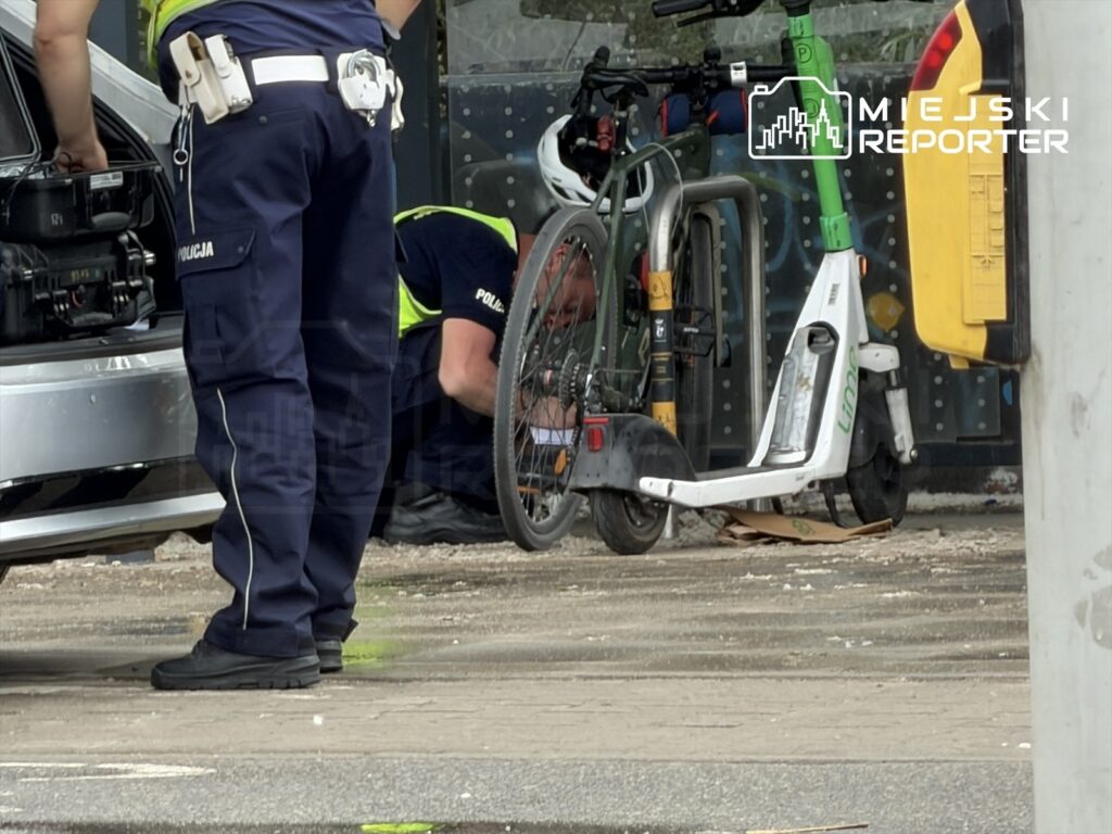 Policjant sprawdzający wnętrze samochodu, podczas gdy inny mężczyzna naprawia rower obok elektrycznej hulajnogi.