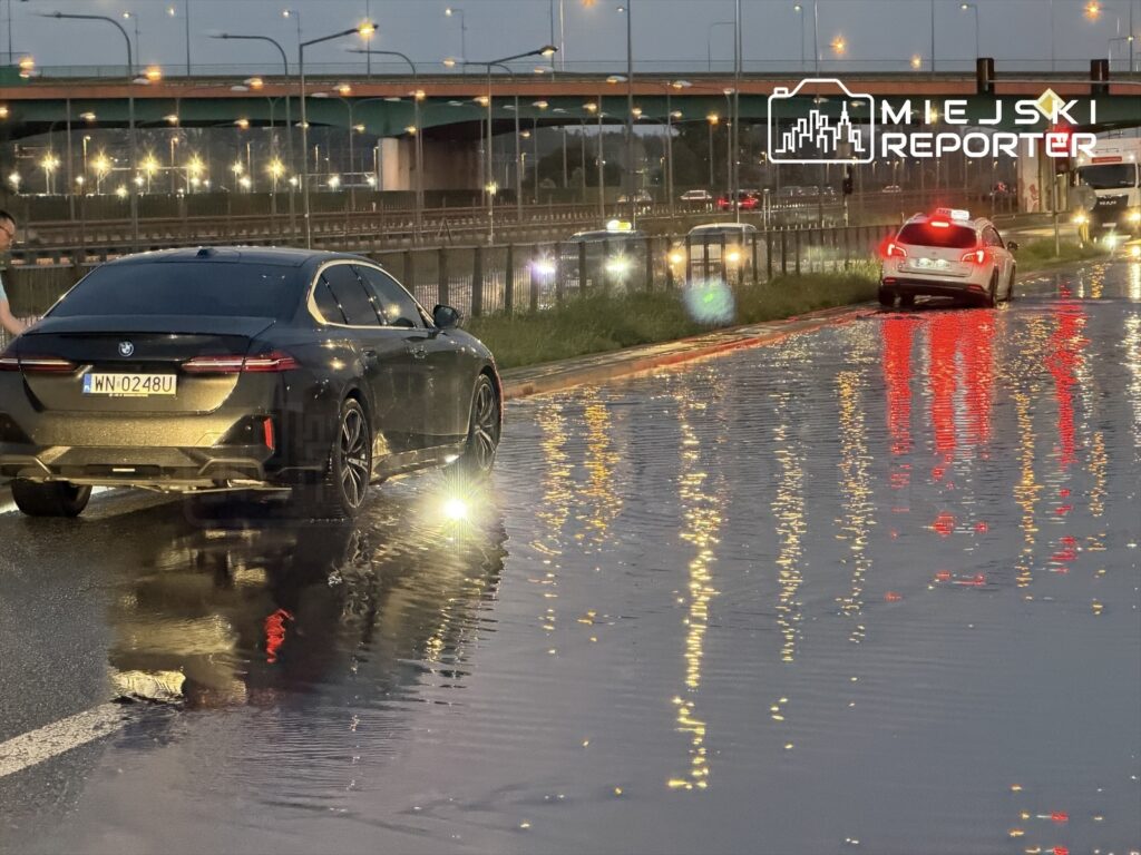 Czarne auto osobowe porusza się po zalanej drodze, odbijając światła z otoczenia. W tle widoczny most.