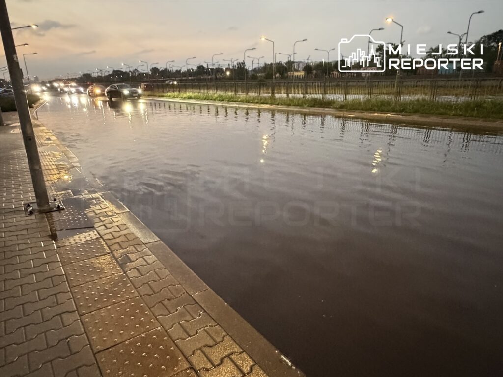Ulica pokryta wodą po intensywnych opadach deszczu, samochody poruszają się wzdłuż zalanego pobocza.