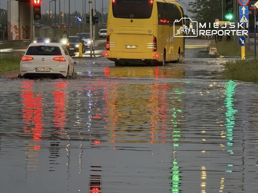 Samochody poruszają się po zalanej drodze, odbijając światła sygnalizacji i otoczenia.