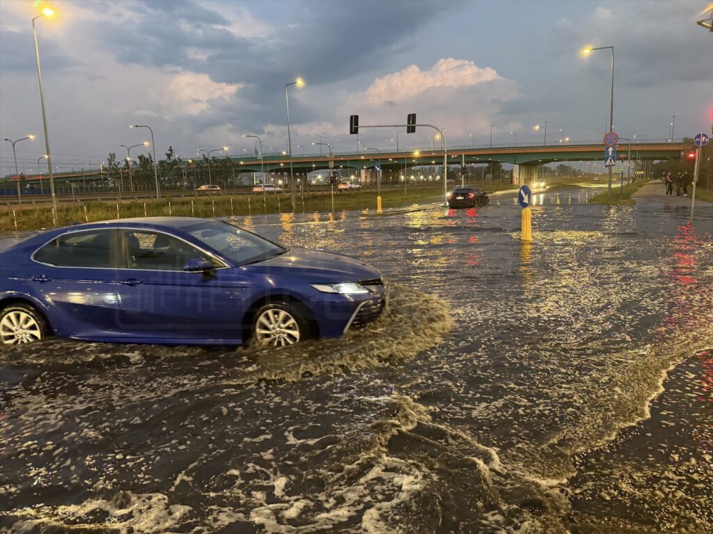 Niebieski samochód osobowy porusza się przez zalaną ulicę w godzinach wieczornych, wokół widoczne są inne pojazdy i latarnie.