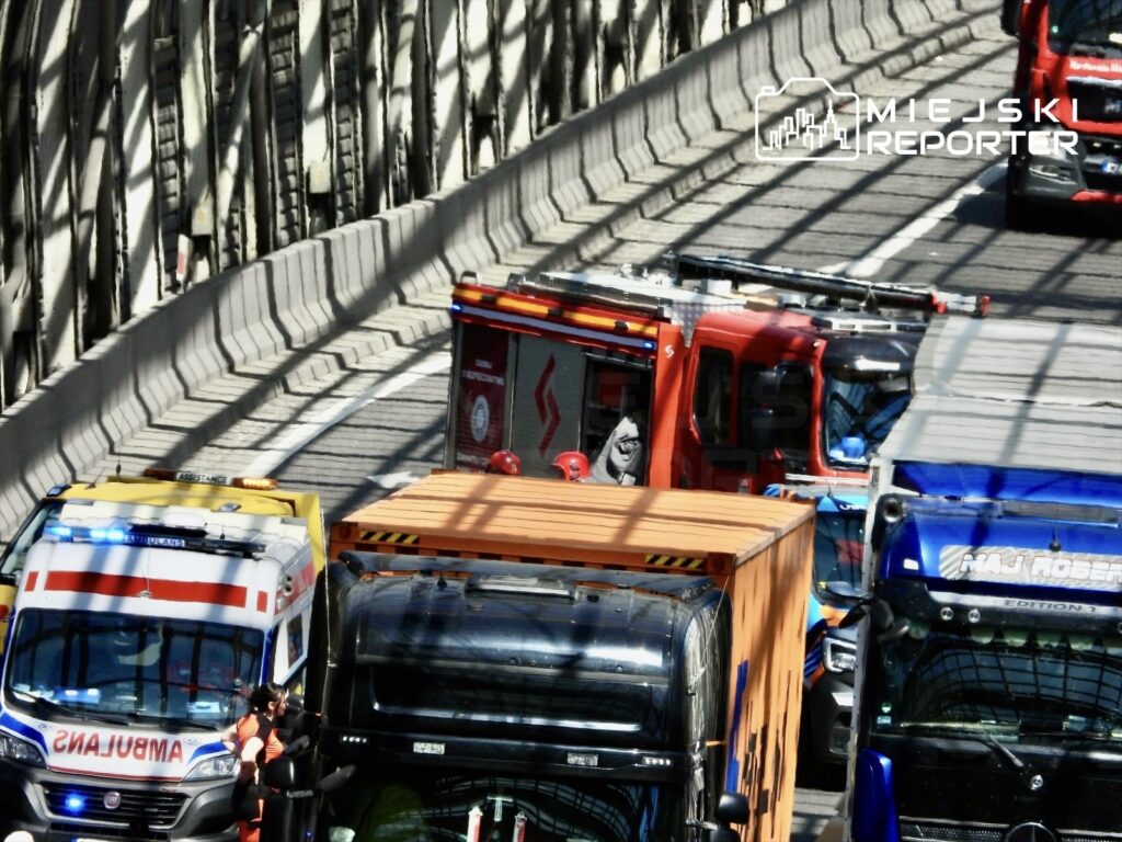 Karetka i wóz strażacki zatrzymane na ruchliwej drodze, obok ciężarówki oraz pojazdów transportowych.