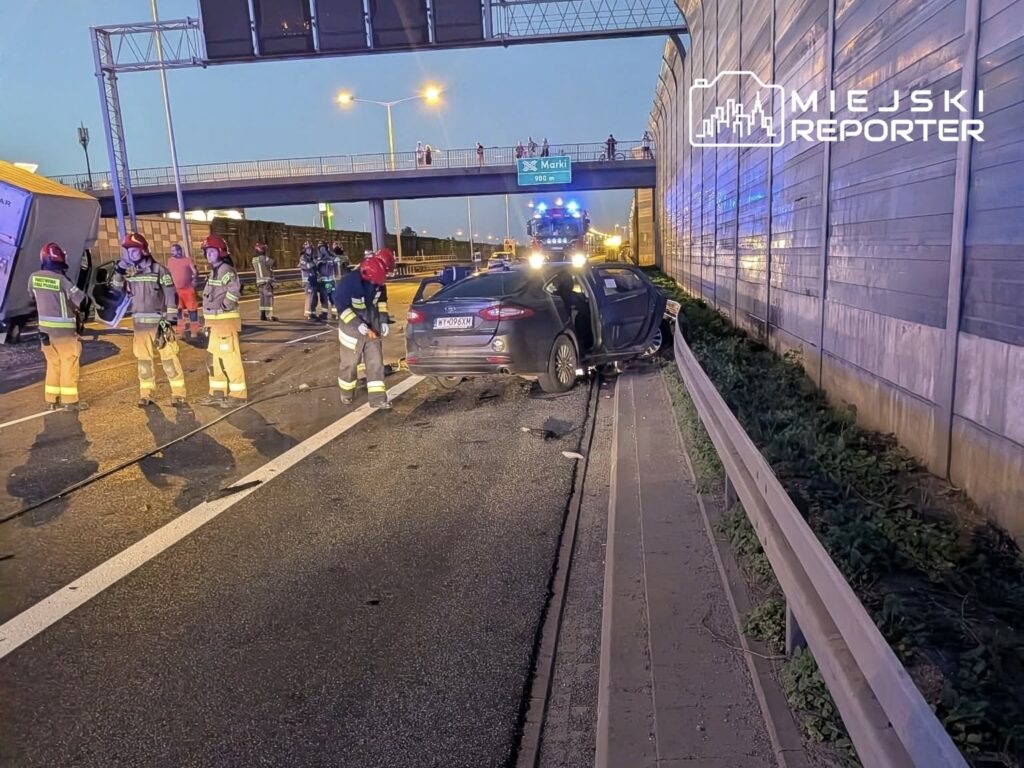 Na drodze leży przewrócony tir, obok grupa strażaków pracujących przy usuwaniu skutków wypadku. W tle widać uszkodzony samochód osobowy.