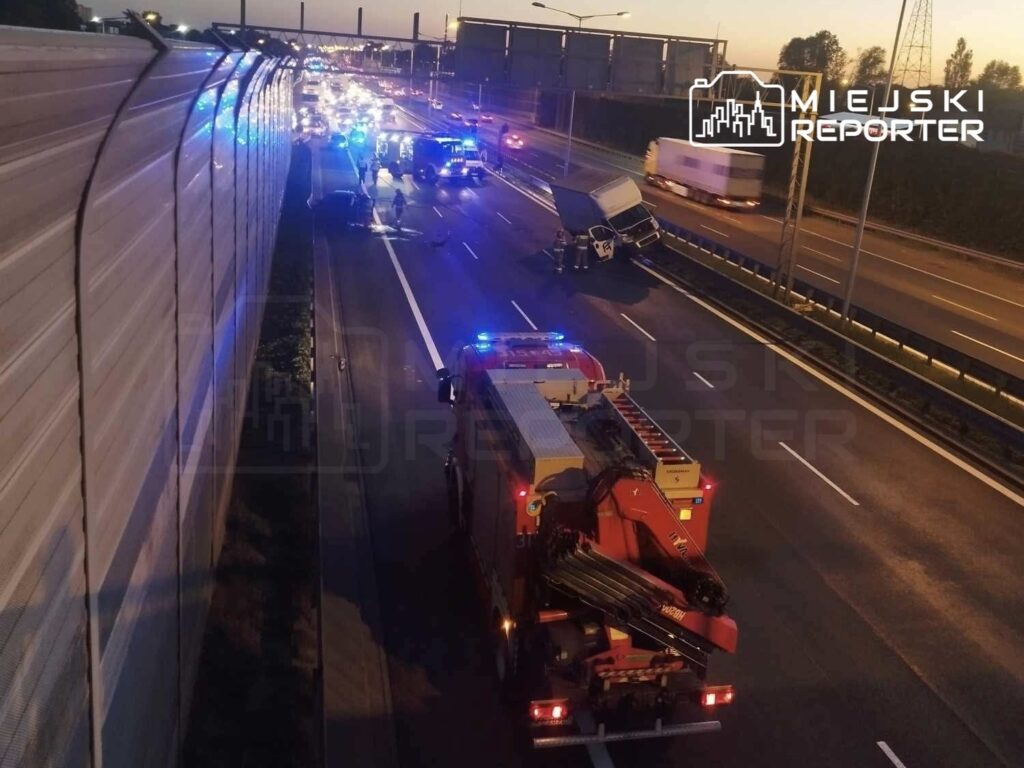 Na autostradzie z włączonymi sygnałami świetlnymi stoją pojazdy straży pożarnej i kilka samochodów, w tym przewrócony dostawczy.