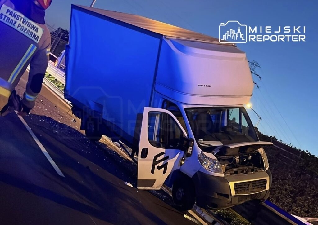Strażak w mundurze klęczy obok przewróconego dostawczego samochodu ciężarowego na jezdni.