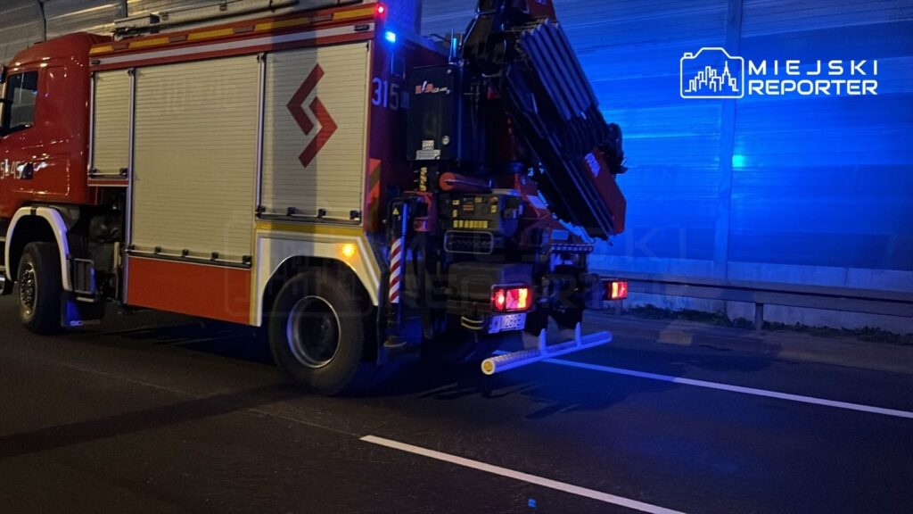 Czerwony wóz strażacki z włączonymi sygnałami świetlnymi stoi na drodze, w tle niebieskie oświetlenie.