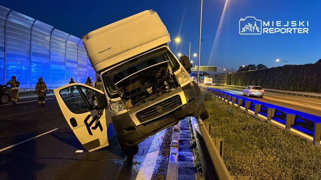 Samochód dostawczy stoi na skraju drogi, z otwartą maską, obok interweniują strażacy w mundurach.