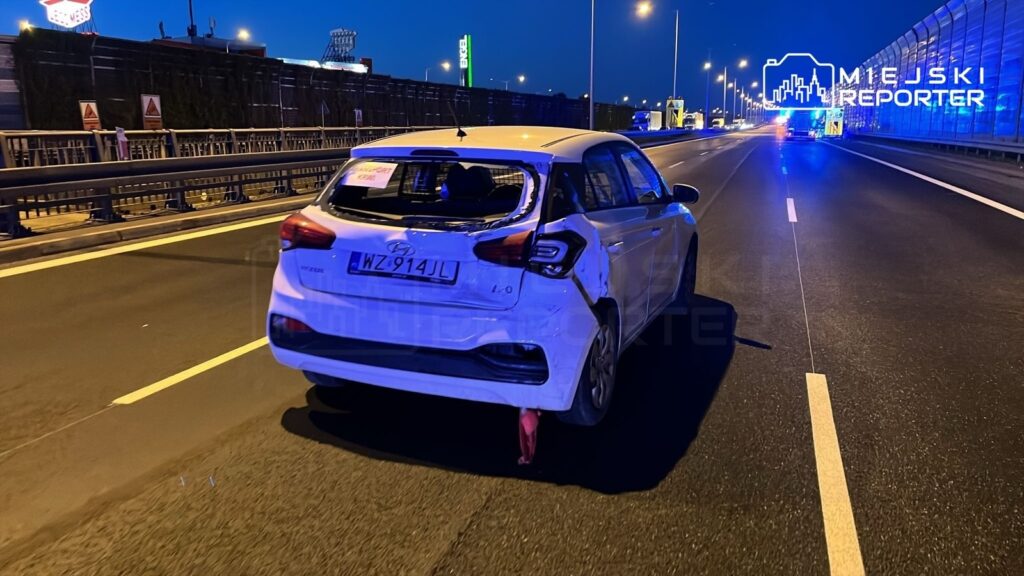 Uszkodzony biały samochód osobowy stoi na autostradzie w nocy, z widocznymi uszkodzeniami tylnej części.