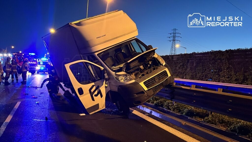 Zniszczony samochód dostawczy stoi na boku przy drodze, otoczony przez strażaków i pojazdy ratunkowe w nocy.