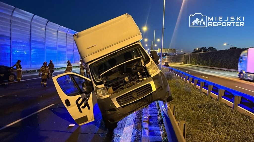 W nocy, na autostradzie, dostawczy samochód stoi na barierkach, a w tle widać strażaków i policyjne światła.