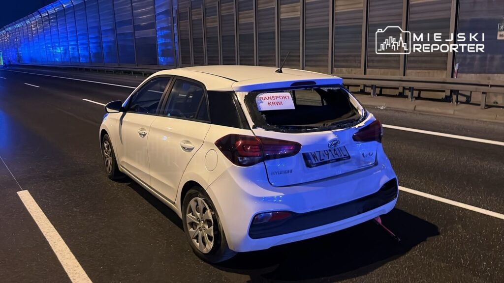 Biały samochód osobowy z uszkodzoną tylną szybą zatrzymany na drodze, z widocznym napisem „TRANSPORT KRWI” na szybie.