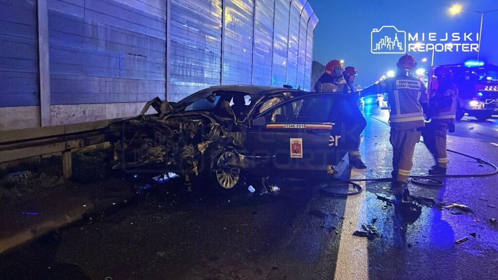 Uszkodzony samochód osobowy stoi na drodze, obok niego pracują strażacy w mundurach, w tle widoczne niebieskie światła.