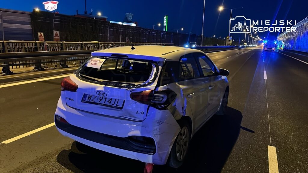 Uszkodzony samochód osobowy zaparkowany na drodze z widocznym napisem "TRANSPORT KRWI". W tle nocne oświetlenie.