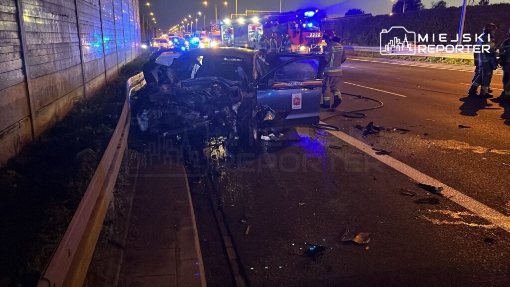 Zniszczony samochód osobowy stoi na poboczu drogi, a obok pracują strażacy w mundurach. W tle widać pojazdy ratunkowe.