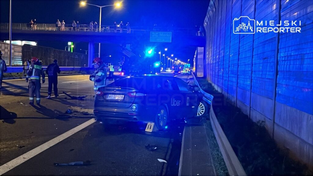Na drodze stoi rozbity samochód Uber, obok którego pracują strażacy w hełmach, a w tle widać sygnalizację świetlną.