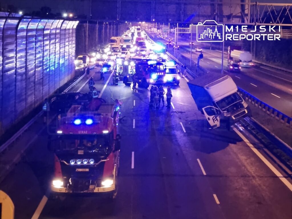 Na drodze stoi przewrócony samochód dostawczy, a w tle widać pojazdy ratunkowe z włączonymi sygnałami świetlnymi.