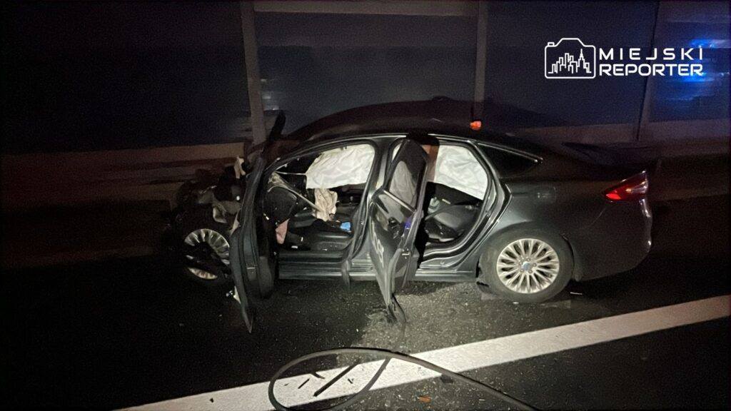 Uszkodzony samochód osobowy z otwartymi drzwiami stoi na jezdni, obok leży przewód. W tle ciemna sceneria nocna.