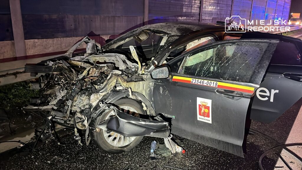 Zniszczony samochód osobowy leży na jezdni, z widocznymi uszkodzeniami przodu i boku, w nocy.