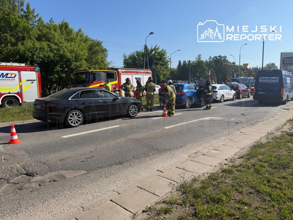Grupa strażaków udziela pomocy poszkodowanym w wypadku drogowym na ruchliwej ulicy, obok zaparkowanych pojazdów.