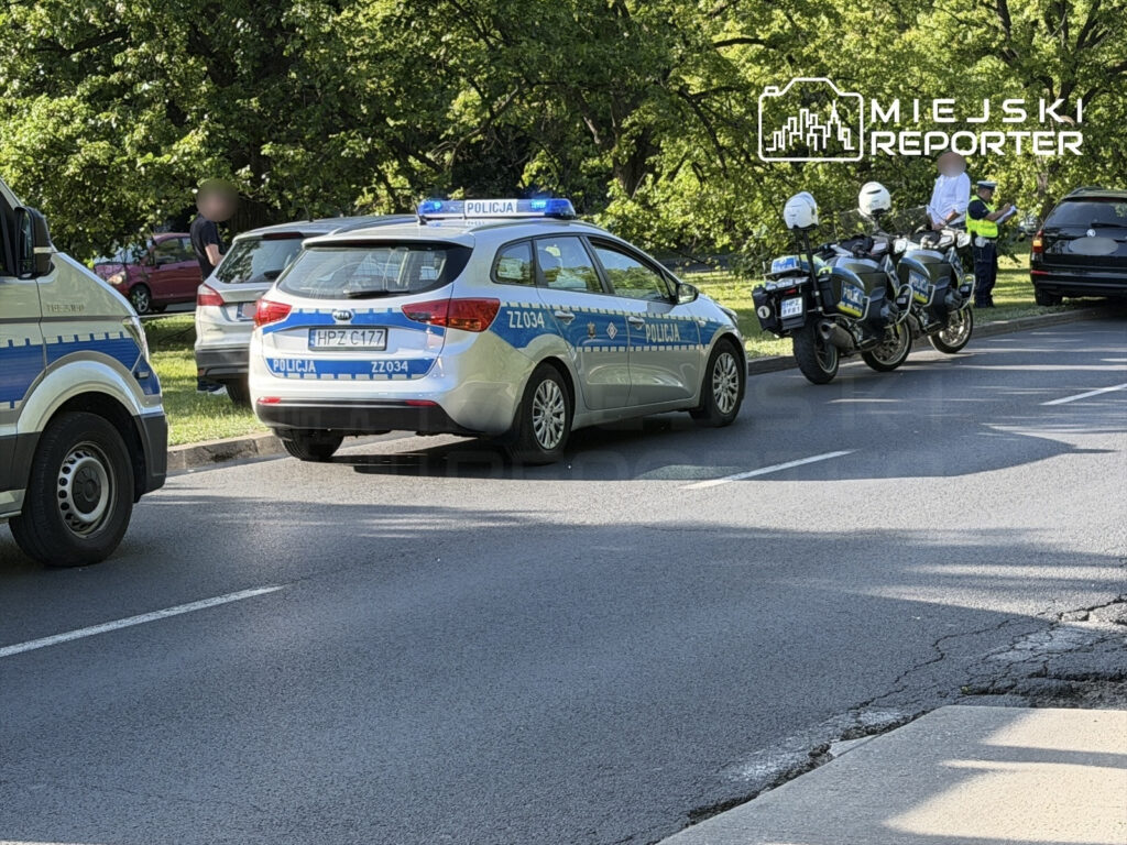Radiowóz policyjny zatrzymany na drodze, obok motocykliści w mundurach, a w tle mężczyzna stojący przy samochodzie.