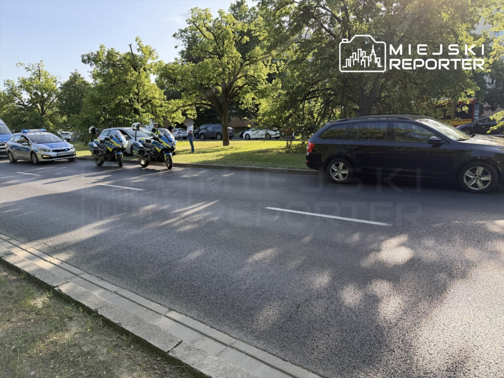 Policyjny radiowóz i motocykle zaparkowane na ulicy w otoczeniu zieleni, obok stojący samochód osobowy.