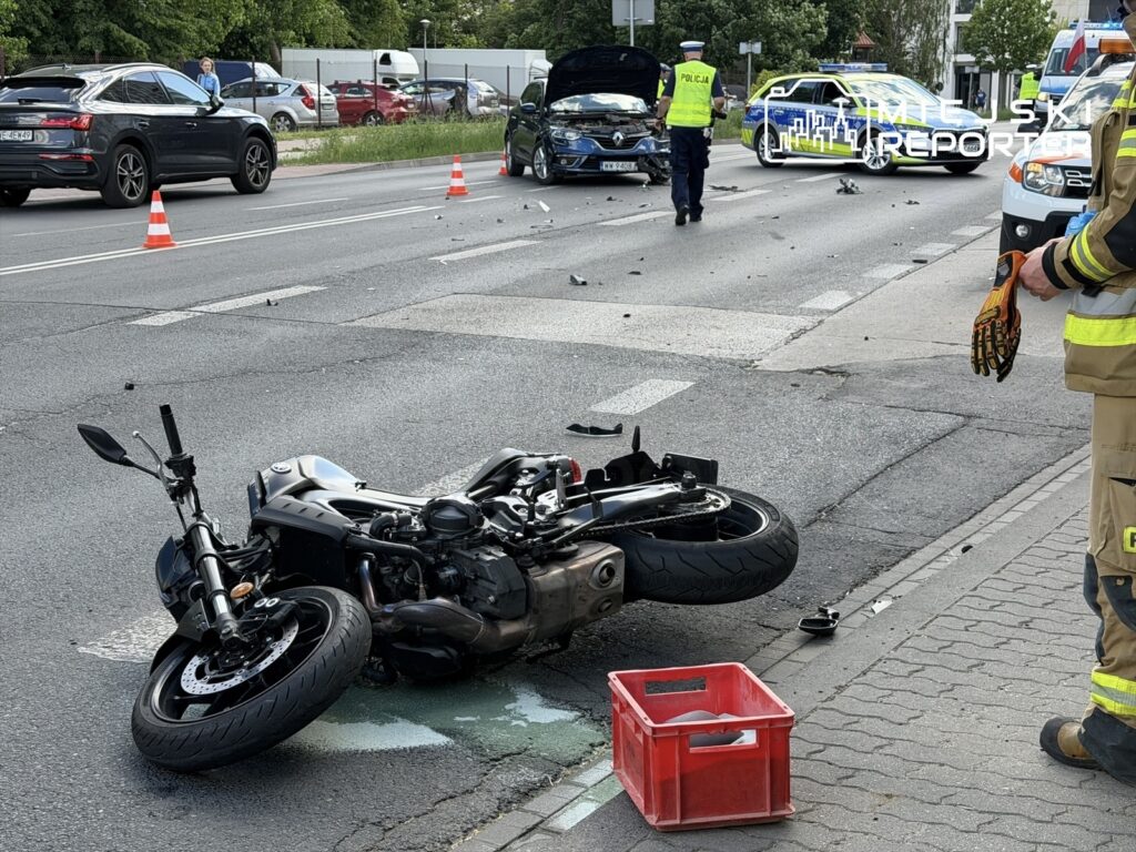 Motocykl leży na jezdni obok rozbitego samochodu, a policjanci i strażacy zabezpieczają miejsce wypadku.