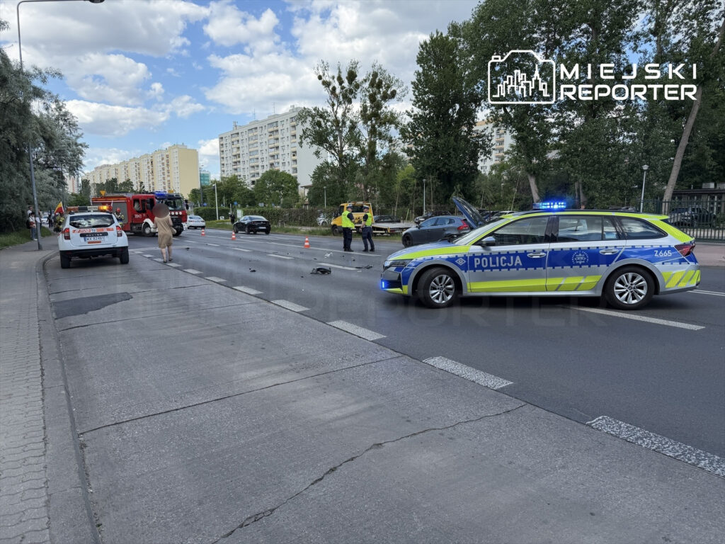 Na jezdni widać wypadek drogowy, w którym uczestniczy radiowóz policji oraz straż pożarna. Na drodze leżą fragmenty pojazdów, a w oddali stoją świadkowie zdarzenia.