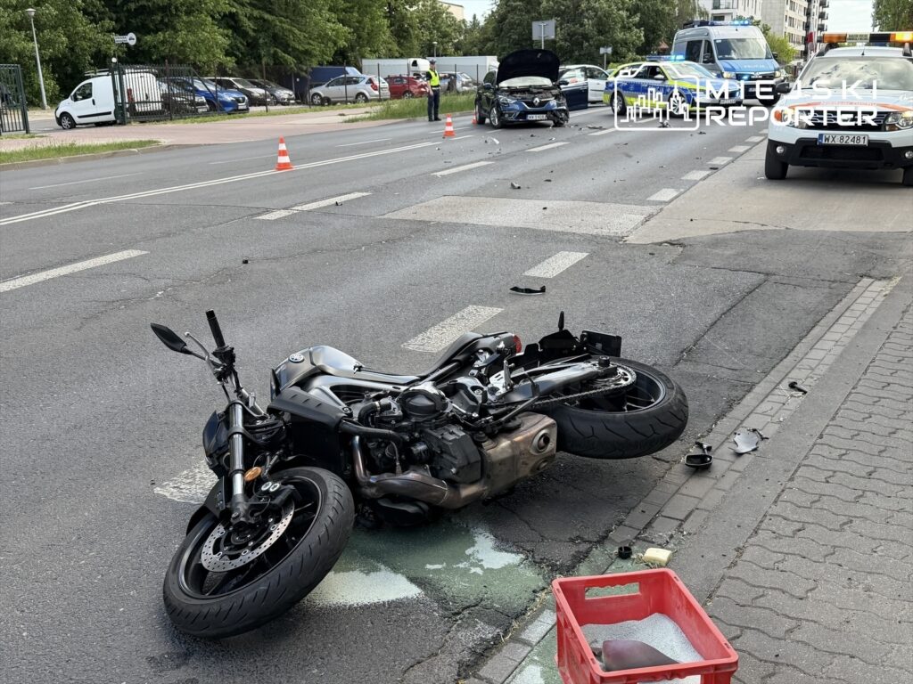 Motocykl leży na boku na jezdni, obok widać uszkodzony samochód osobowy oraz rozbite elementy. W tle stoją radiowozy policyjne.