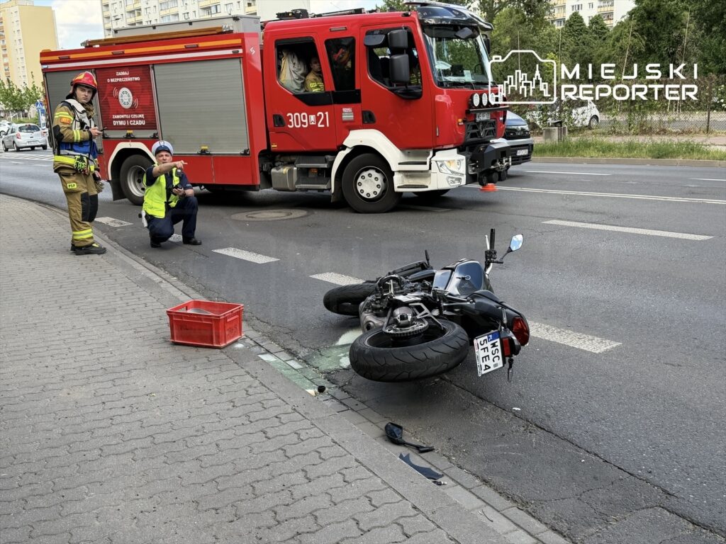 Strażak w mundurze i policjant badają przewrócony motocykl na jezdni, obok stoi czerwona skrzynka.