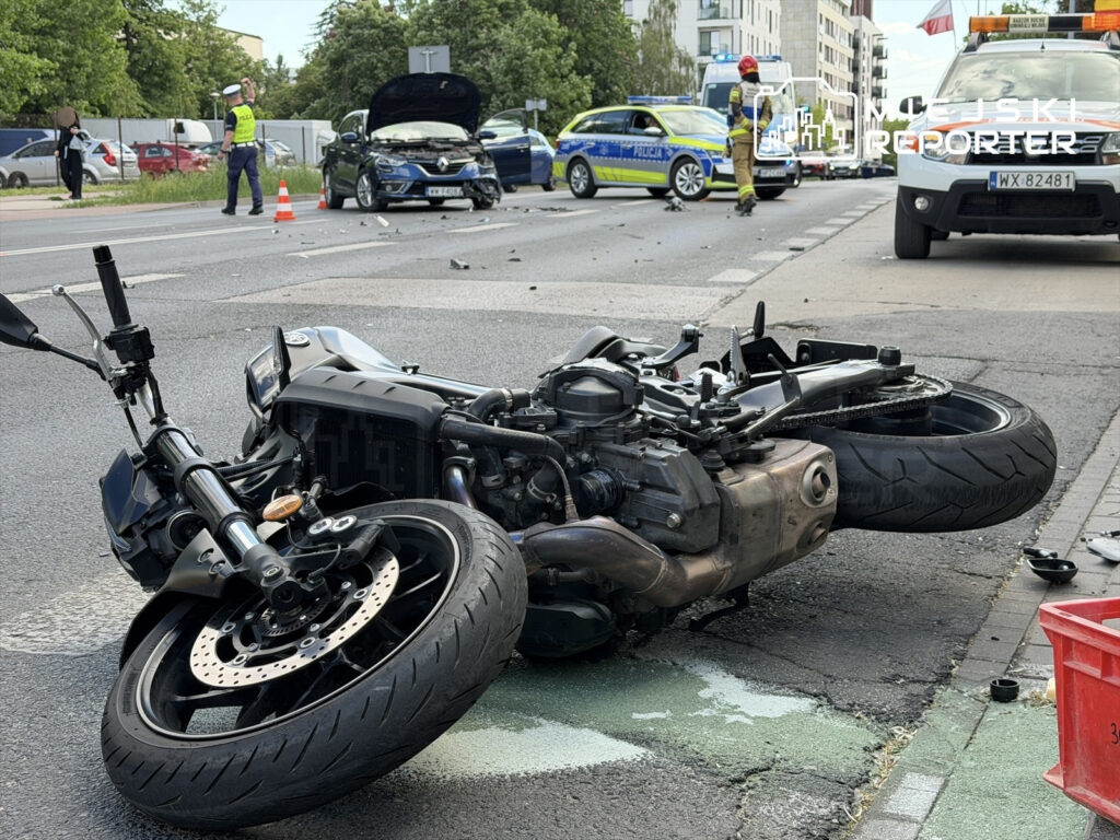 Motocykl leży na boku na jezdni, obok widać uszkodzony samochód oraz policjantów zabezpieczających miejsce zdarzenia.