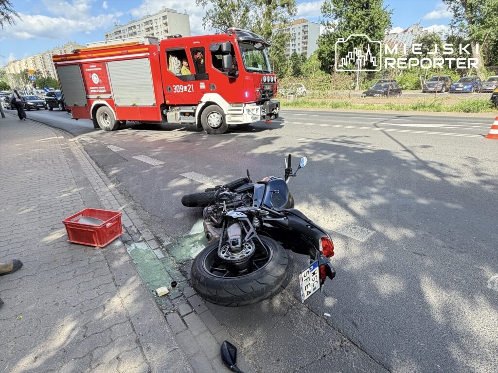 Motocykl leżący na boku obok czerwonego wozu strażackiego na ulicy, obok widoczna czerwona skrzynka.
