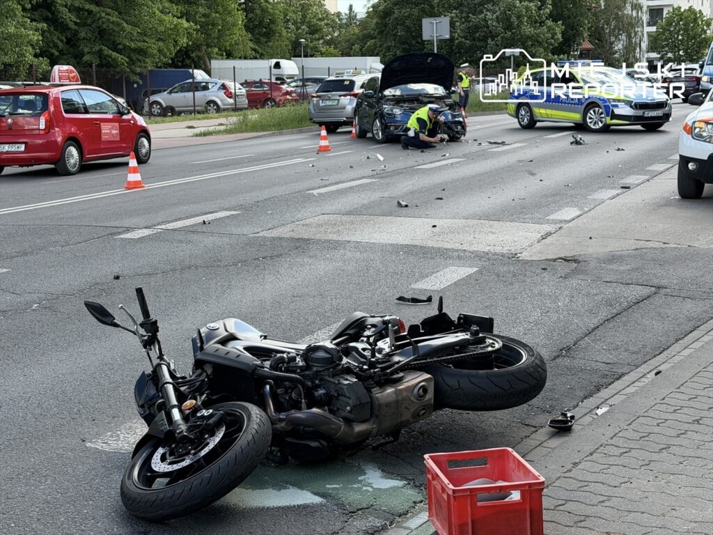 Motocykl leży na boku na jezdni, a obok pracują policjanci przy uszkodzonym samochodzie. W tle widać korki.