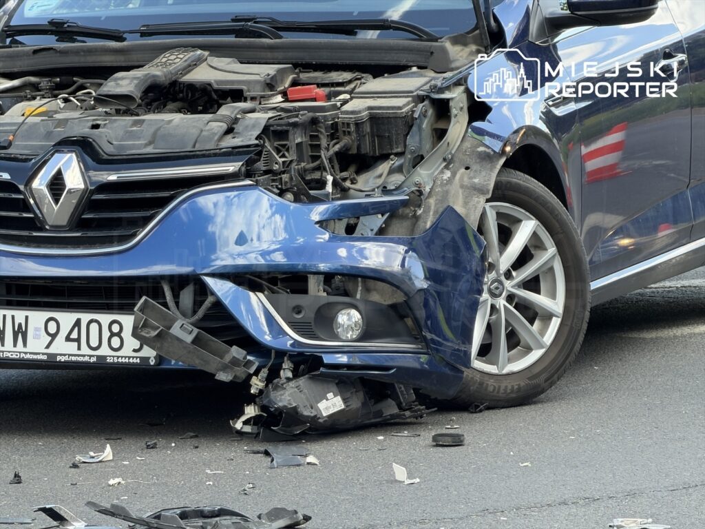 Uszkodzony niebieski samochód osobowy z widoczną uszkodzoną przednią częścią oraz częściami leżącymi na jezdni.