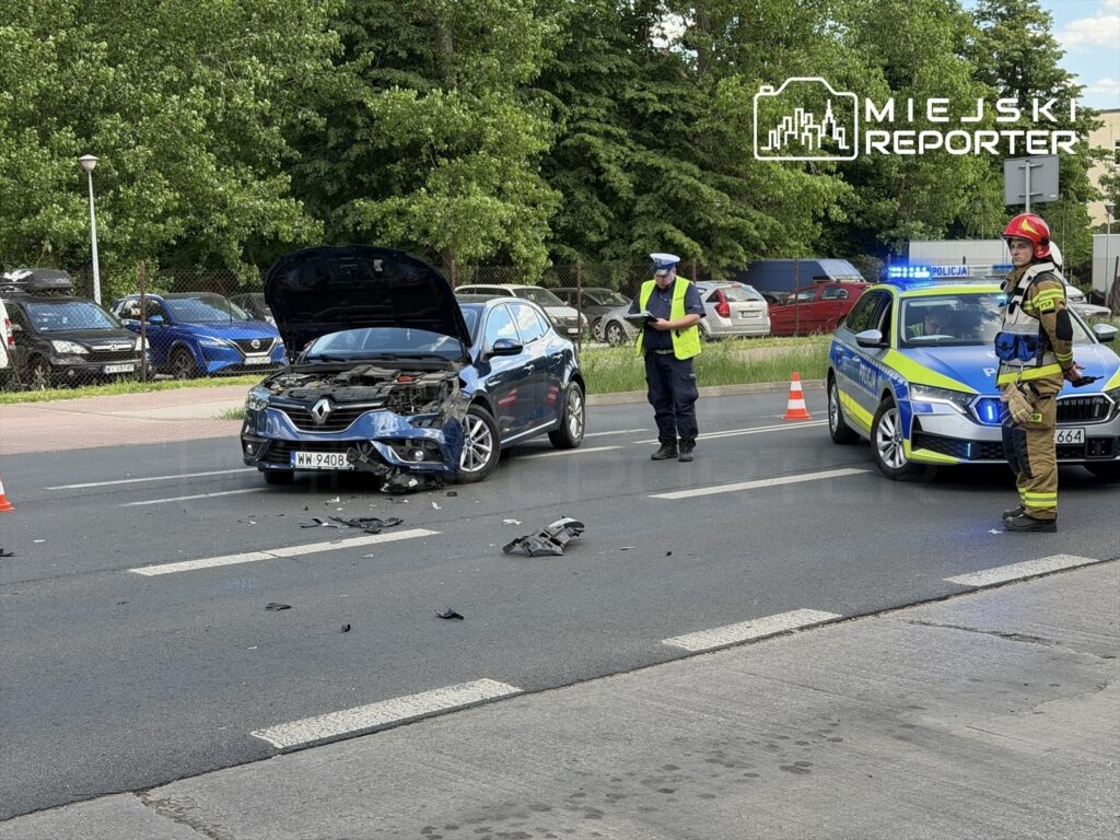 Renault z uszkodzoną maską stoi na jezdni, obok pracują policjant i strażak, a w tle widać inne pojazdy.